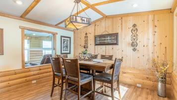 A wooden-paneled dining room with a large table and chairs, a hanging light fixture, and framed pictures on the walls.