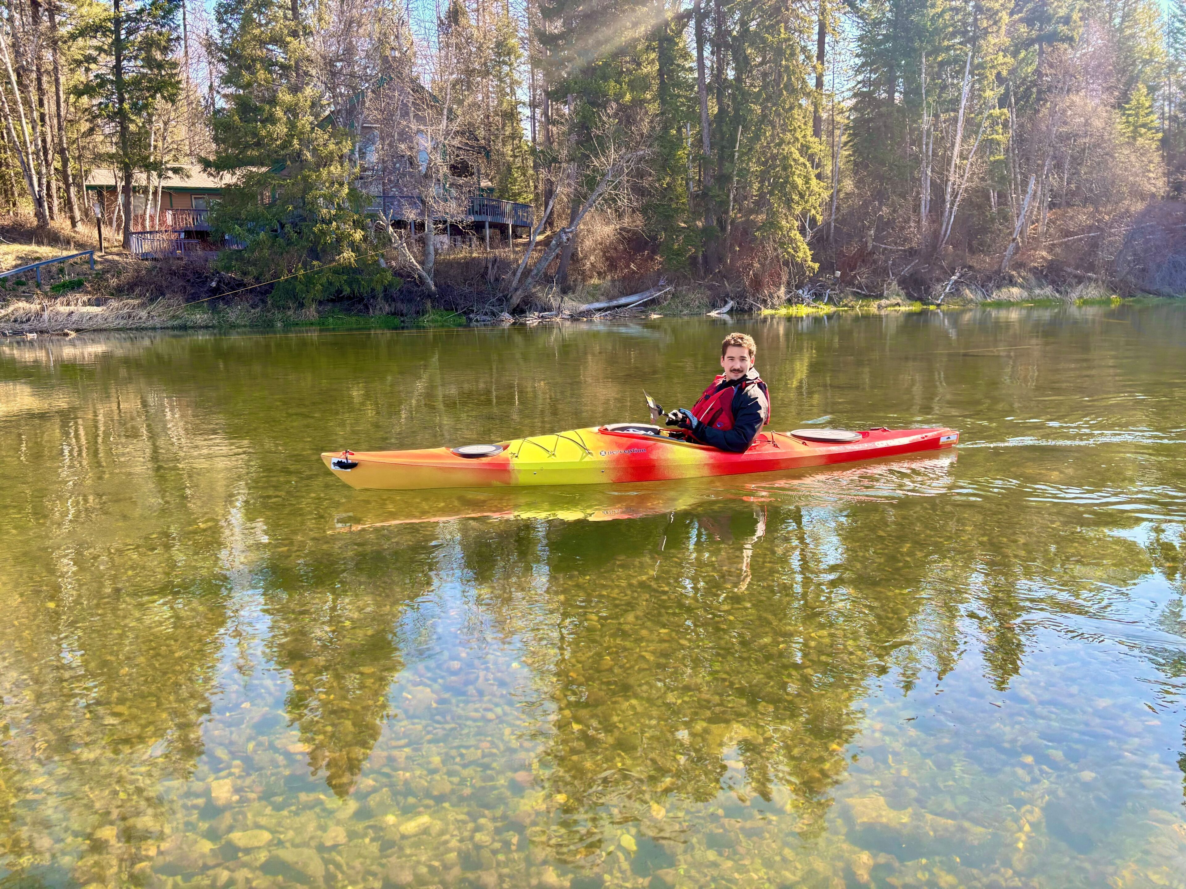 Eine Person sitzt in einem roten Kajak auf einem ruhigen See, umgeben von Bäumen.