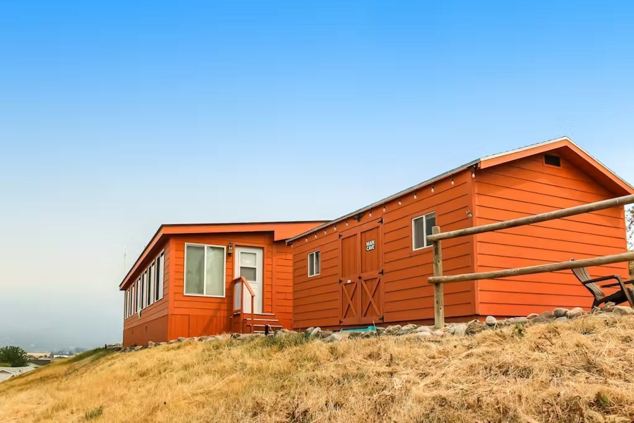 Une cabane en bois rouge, dotée d’une grande fenêtre et d’une porte, située sur une colline verdoyante sous un ciel dégagé.
