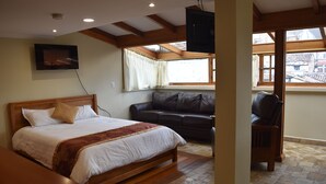 Ein Schlafzimmer mit einem Bett, einem Ledersofa, einem Holzstuhl, einem an der Wand befestigten Fernseher und einem Fenster mit Blick auf Gebäude.