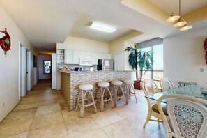 A modern kitchen with white cabinets, a central island with stools, and a dining area with a glass table and wicker chairs. - Shorefront Corner PH, 3BR: Suntide III - Unit 1201 (South Padre Island)