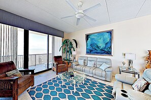 A spacious living room with a large window, a ceiling fan, a sofa, a coffee table, and a painting on the wall. - Shorefront Corner PH, 3BR: Suntide III - Unit 1201 (South Padre Island)