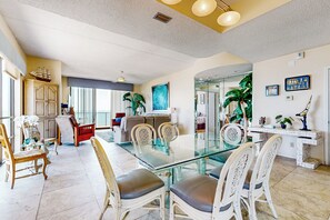 A spacious dining area with a glass table and white chairs, a wooden cabinet, and a large window with curtains. - Shorefront Corner PH, 3BR: Suntide III - Unit 1201 (South Padre Island)
