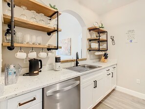 Eine moderne Küche mit weißen Schränken, Edelstahlgeräten und Holzregalen, auf denen Glaswaren und Pflanzen stehen.