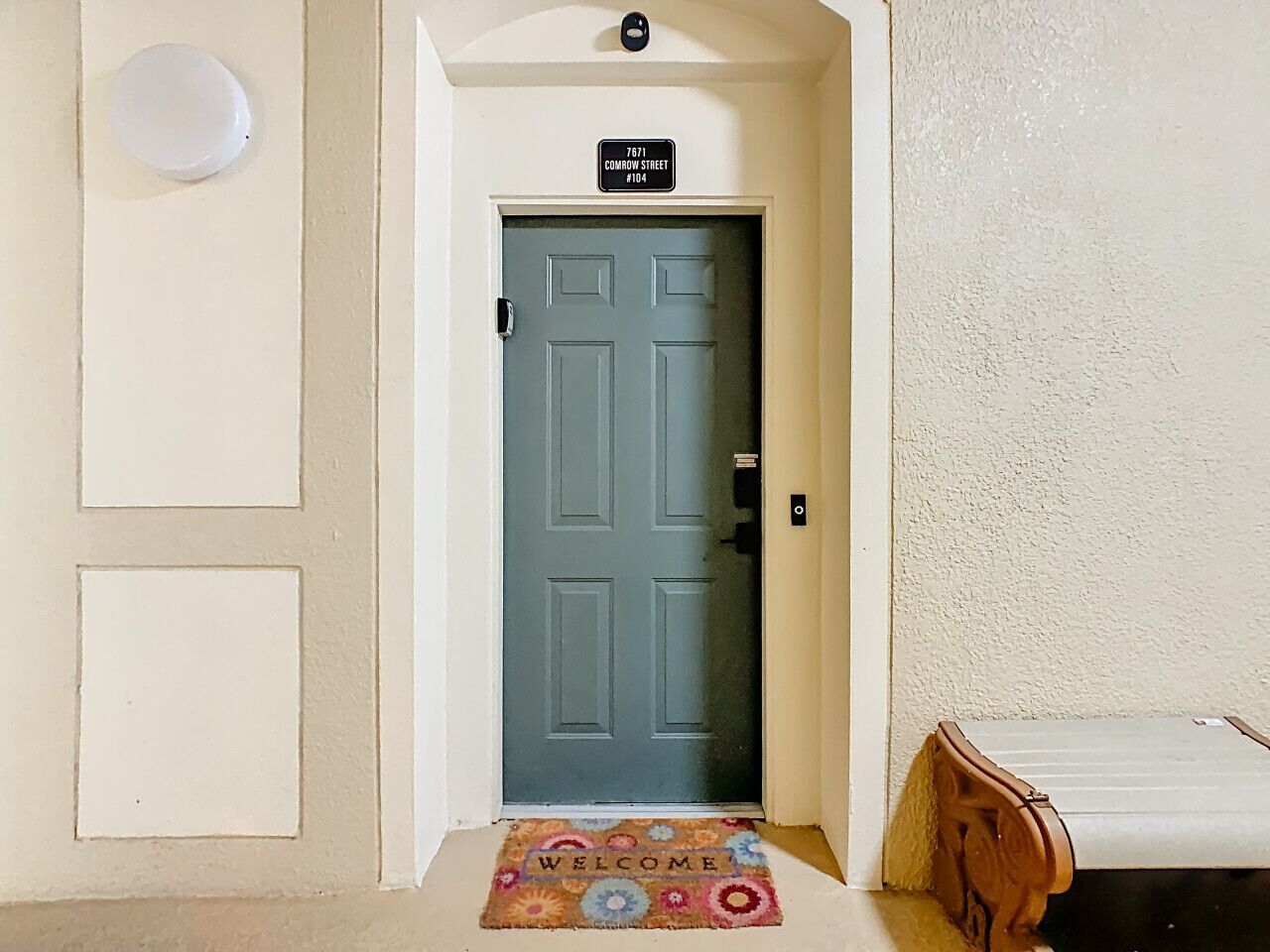 A hallway with a blue door, a welcome mat, and a sign above the door indicating the address.