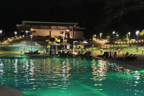 Uma área de piscina à noite, com espreguiçadeiras e guarda-sóis, rodeada por um edifício com varanda coberta.