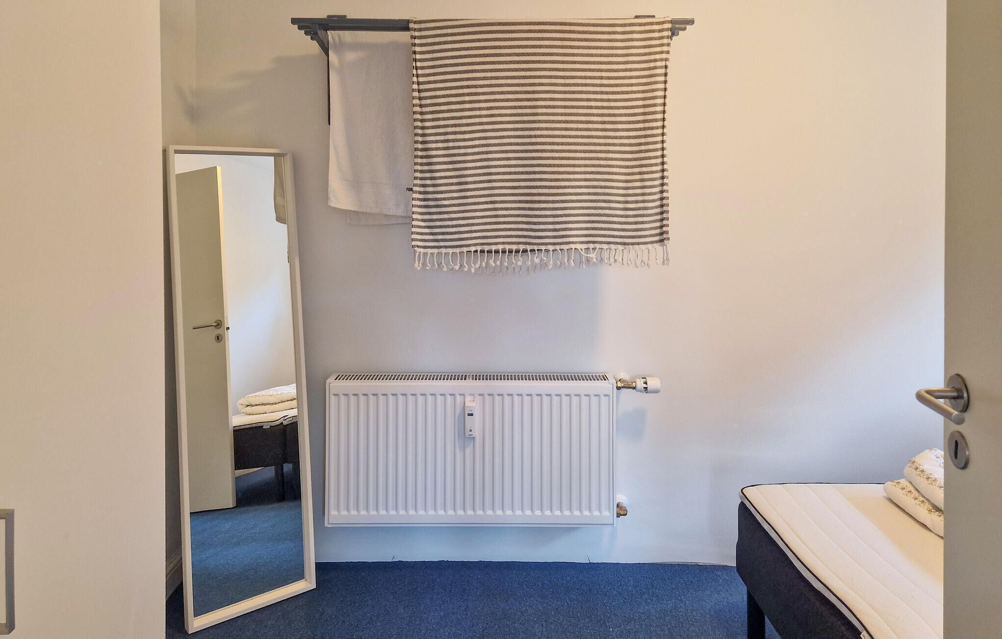 Une chambre avec un radiateur blanc, un lit recouvert d’une couverture beige, une moquette bleue et un miroir qui reflète une partie de la pièce.
