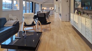 Una sala moderna con una mesa de centro negra, un sillón blanco mullido y un sofá azul. Cuenta con una cocina con armarios blancos y un comedor con una mesa larga y sillas.