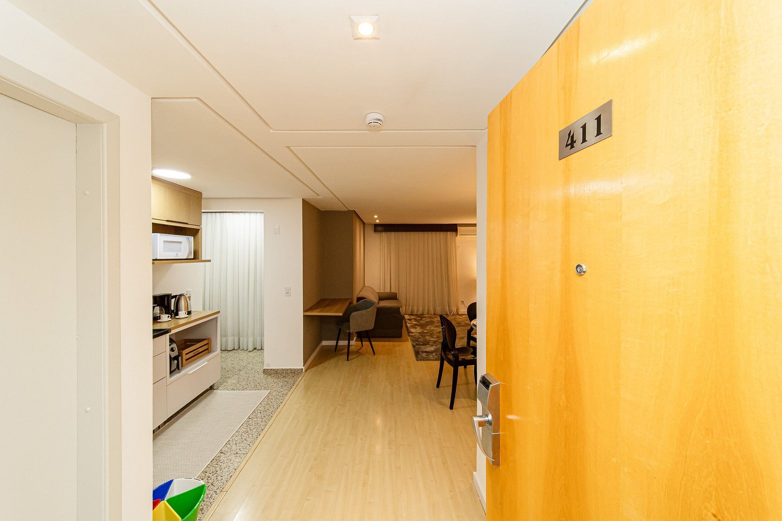 Hotelkamer met een houten deur waarop '411' staat, een kitchenette, een eethoek met tafel en stoelen, en een bank.