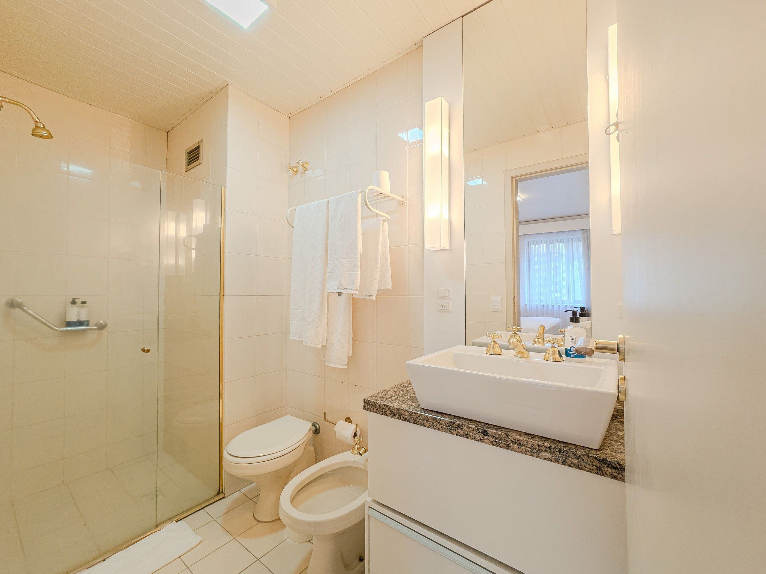 A bathroom with a glass shower enclosure, a toilet, a bidet, and a sink with gold faucets.