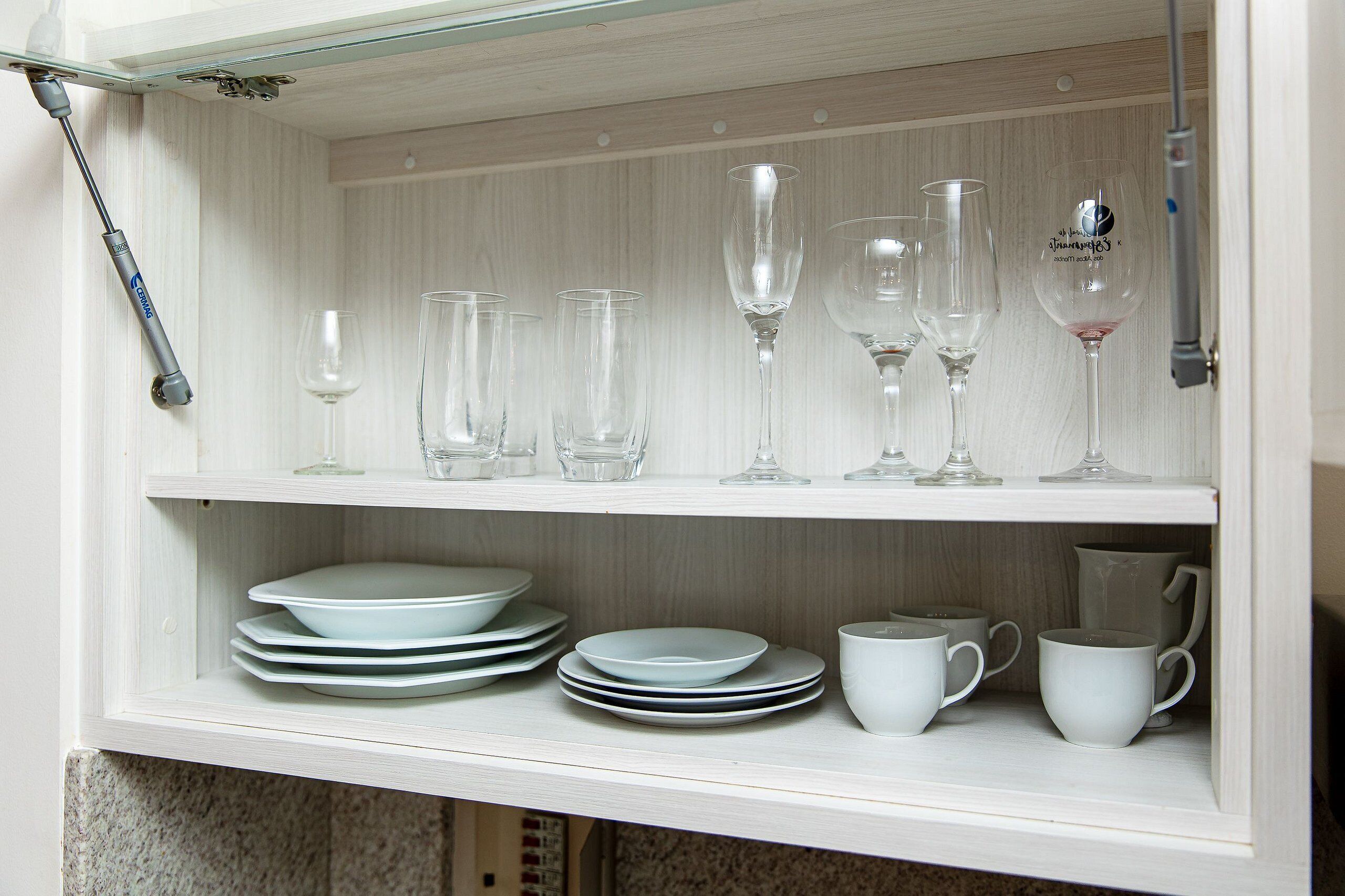 A white cabinet with shelves containing clear wine glasses, plates, and cups.