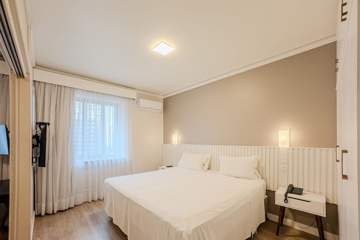 A hotel room with a large bed, a bedside table with a phone, a window with curtains, and an air conditioning unit.
