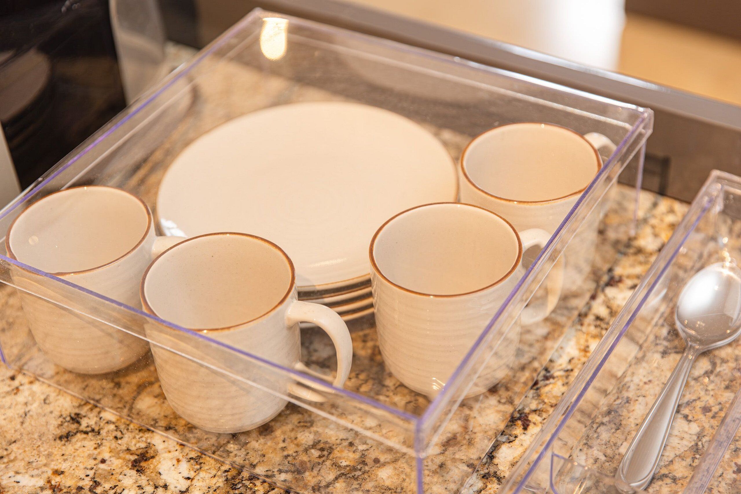 Un organizador de plástico transparente sobre una encimera de granito con platos y tazas blancos.