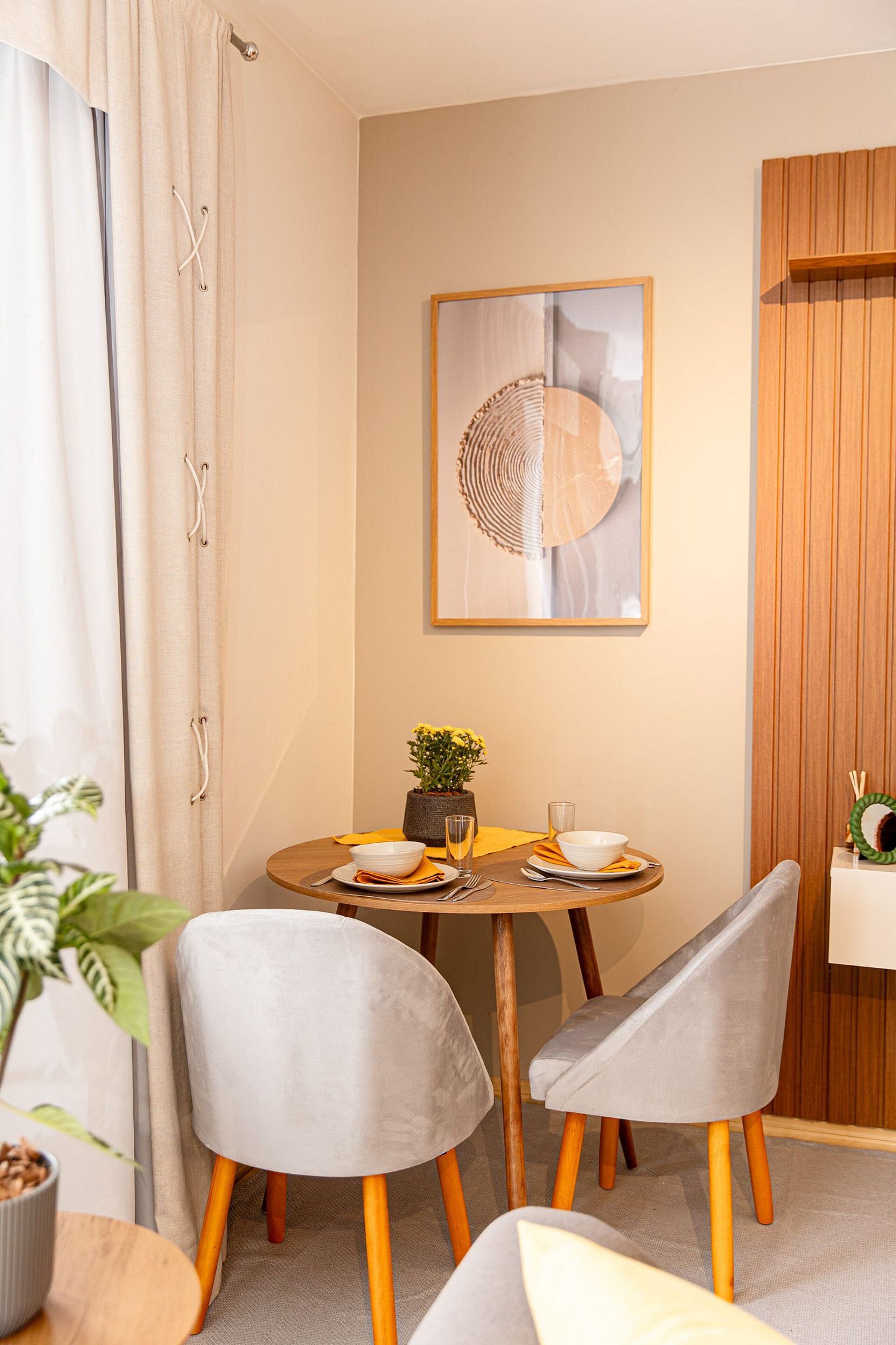 A dining area with a round table, two grey chairs, a potted plant, and a framed artwork on the wall.