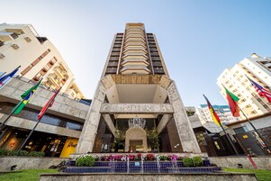 Um edifício alto com entrada central, várias varandas e bandeiras.