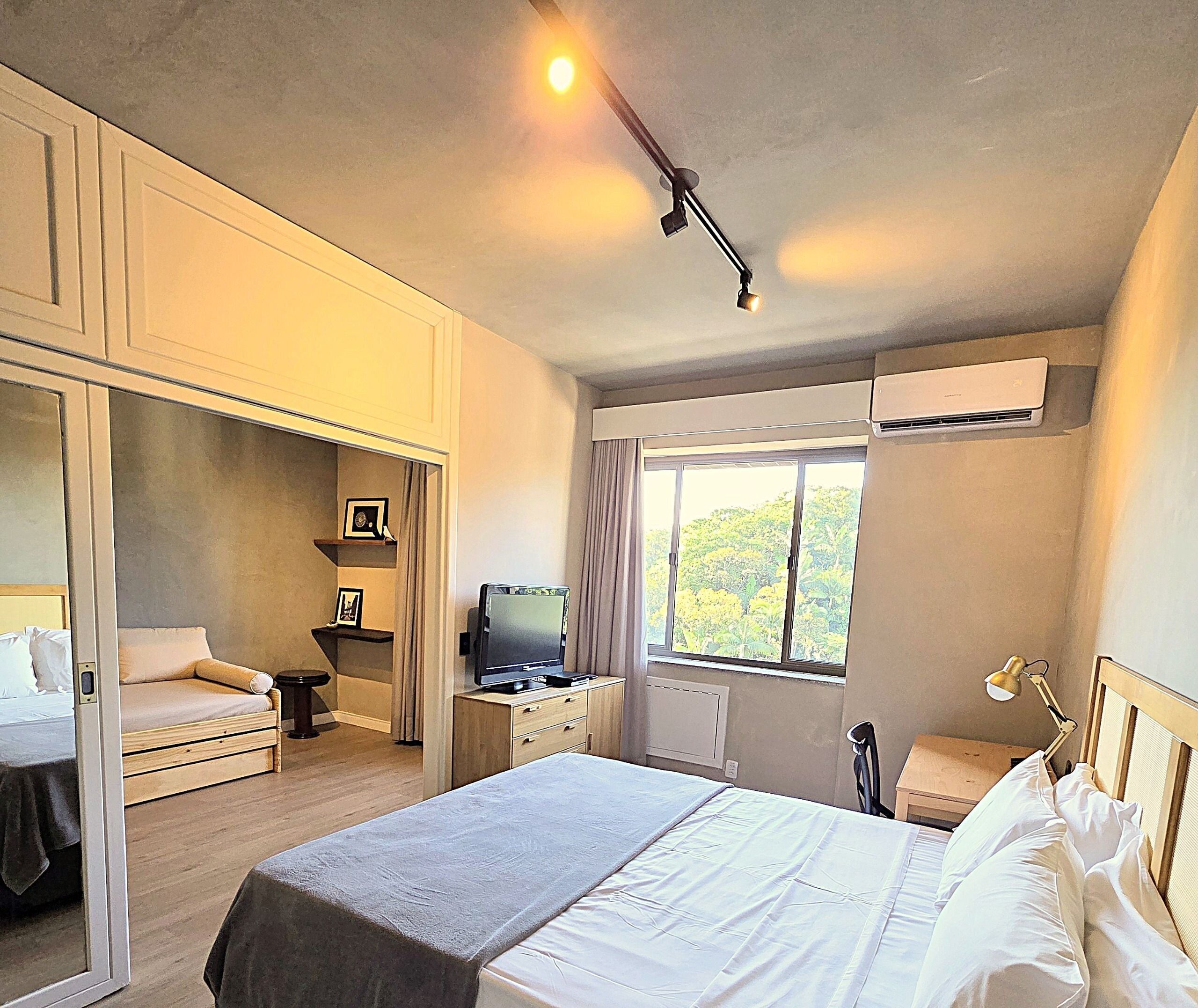 Une chambre à coucher comprenant un lit, un bureau, une télévision, une armoire et une fenêtre donnant sur des arbres.