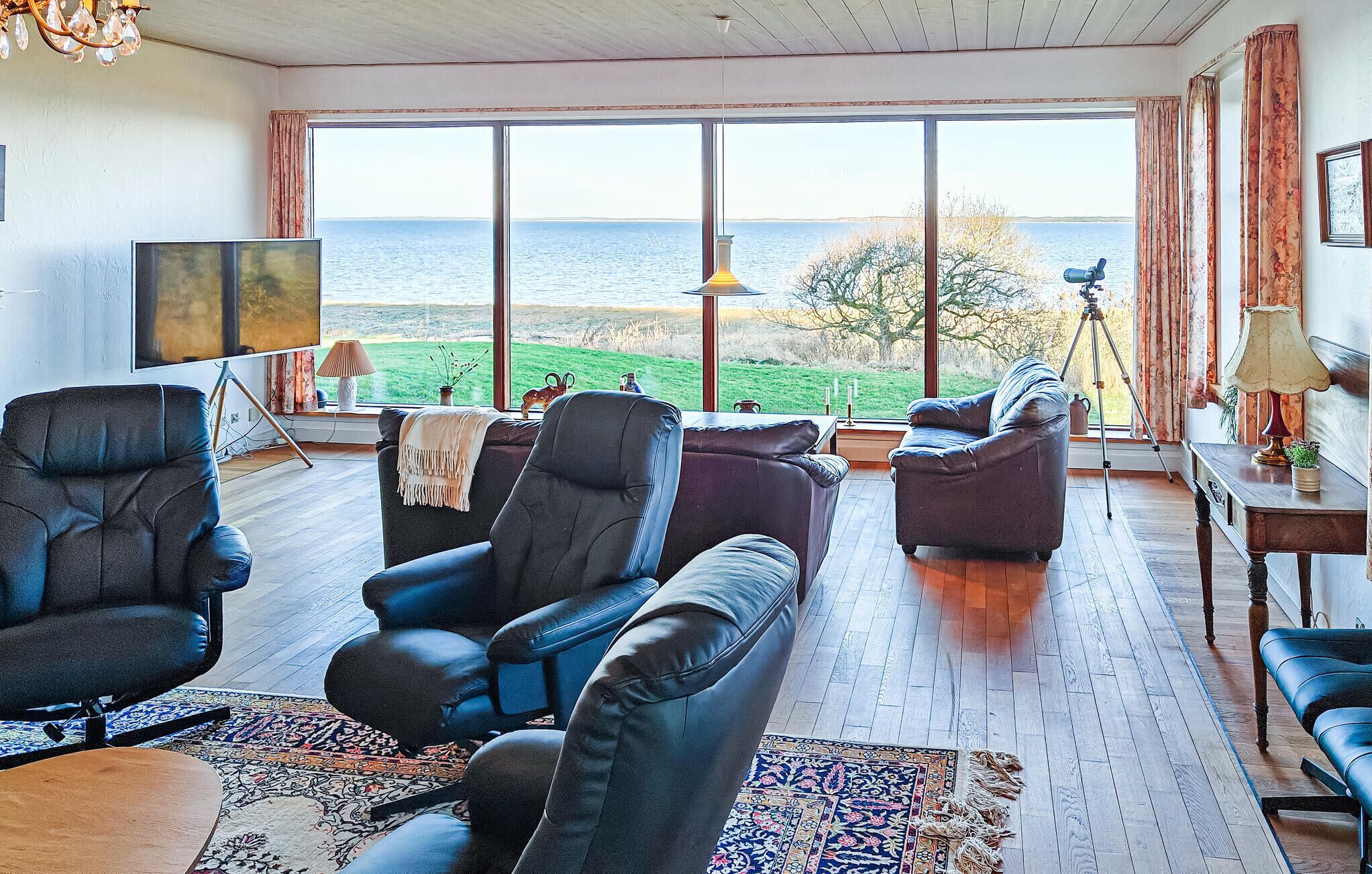 Un vaste salon doté de grandes fenêtres offrant une vue sur la mer, de canapés en cuir, d’un téléviseur et d’un trépied avec un appareil photo.