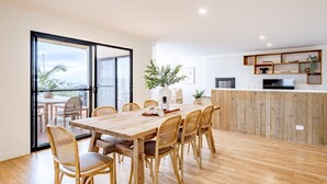A modern dining area with a wooden table and chairs, a potted plant, and a view through a glass door to an outdoor balcony. - Curragudde Cove - Pambula Beach (Pambula Beach)