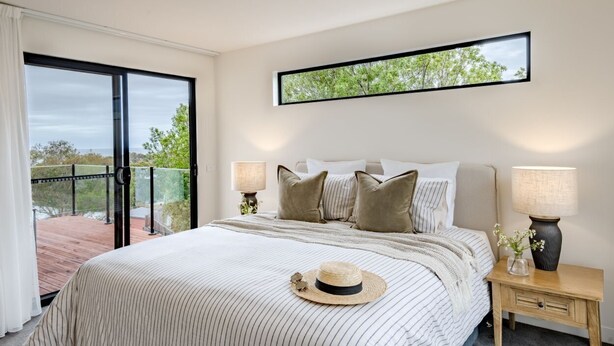 A bedroom with a large bed, two bedside lamps, a nightstand, a window with a view of trees, and a balcony. - Curragudde Cove - Pambula Beach (Pambula Beach)