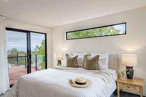 Un dormitorio con una cama grande, dos lámparas de noche, una mesita de noche, una ventana con vistas a árboles y un balcón.