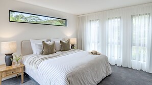 A bedroom with a large bed, two bedside tables, a wall-mounted TV, and sheer curtains. - Curragudde Cove - Pambula Beach (Pambula Beach)