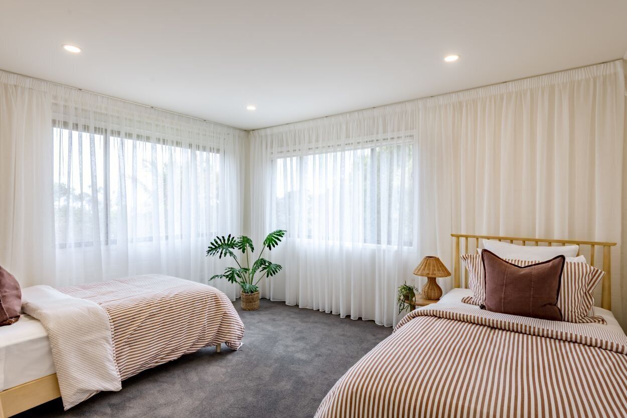 A bedroom with two beds, white curtains, a potted plant, and a bedside lamp.