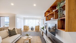 A modern living room with a sofa, two armchairs, a coffee table, and a TV. There are wooden shelves with plants and books. - Curragudde Cove - Pambula Beach (Pambula Beach)