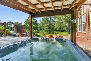 A hot tub with a wooden deck, surrounded by outdoor furniture and a wooden cabin. - Waterfront Bourbon Barrel Cabin (Indianapolis)