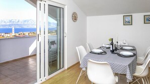 A dining area with a table set for four, white chairs, a clock on the wall, and a view of the sea and mountains through a sliding glass door.