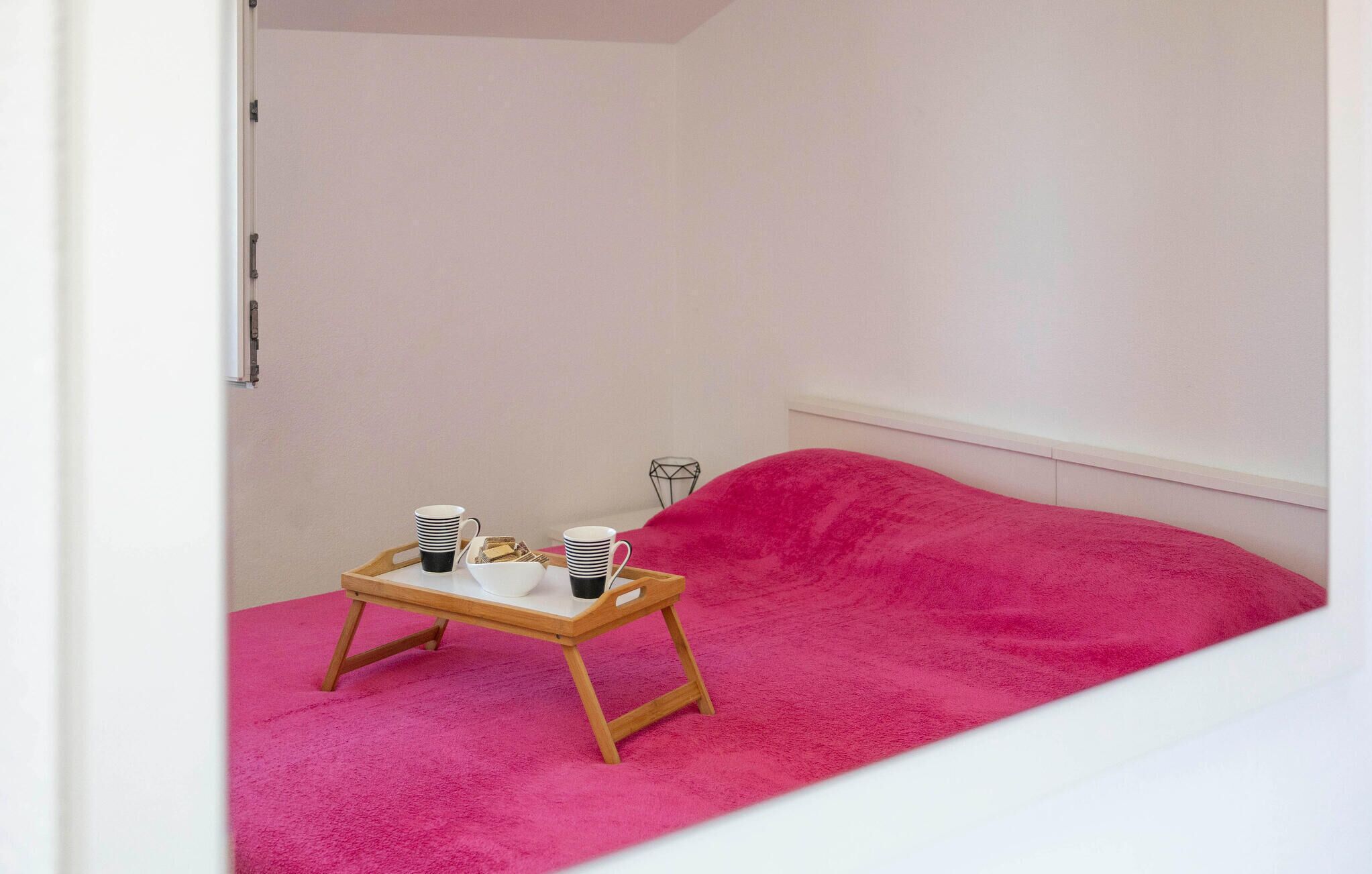 A small room with a pink carpet, a wooden table with cups and a bowl, and a bed with a pink blanket.