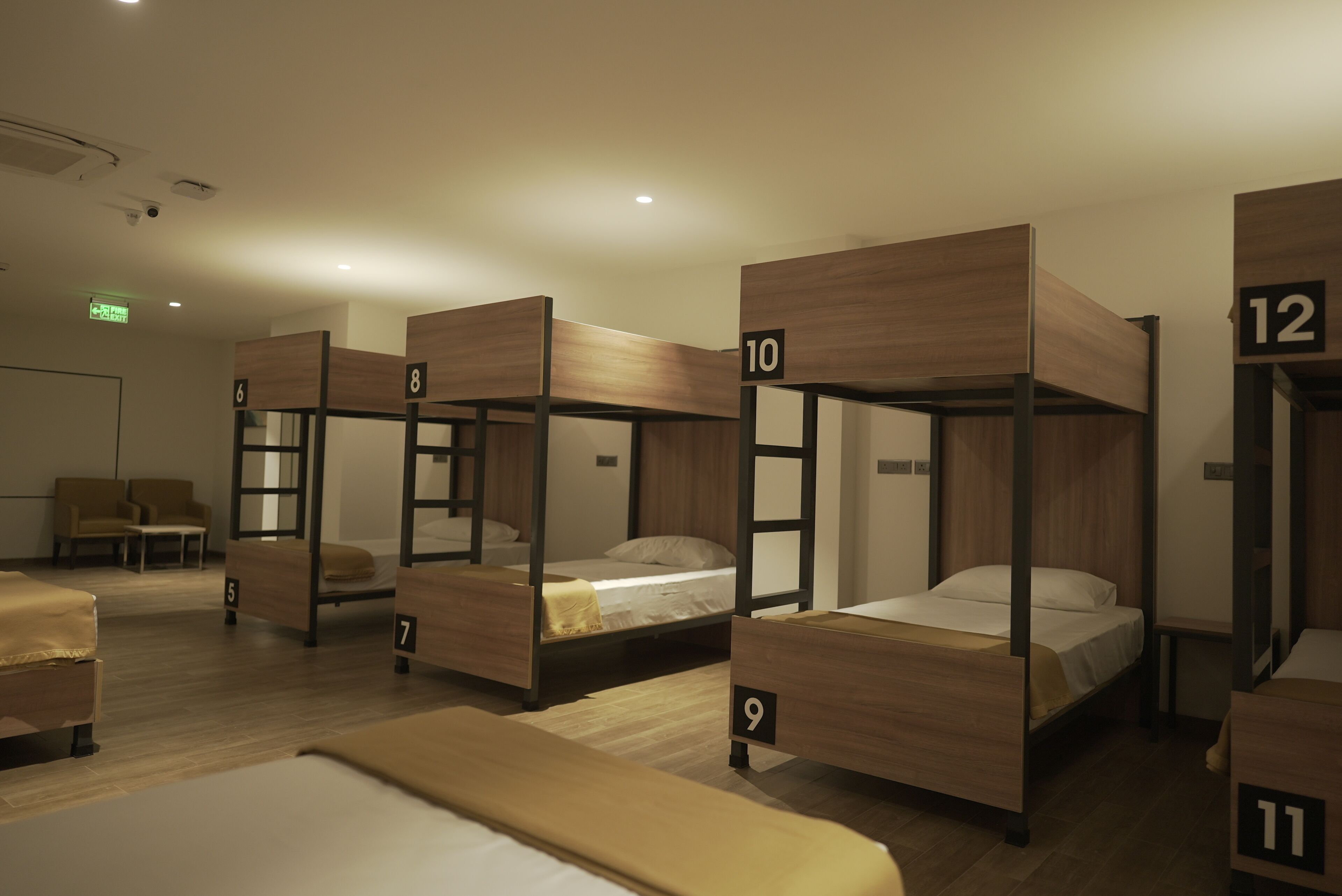 A hotel room with bunk beds numbered 5 to 12, wooden flooring, and a small seating area with chairs.