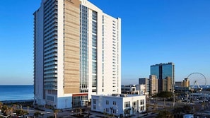 Un hotel moderno de gran altura, situado cerca de una zona costera con una noria en el fondo.