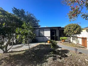 A residential property with a small house, a paved driveway, and a garden area with potted plants and a tree.