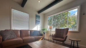 Una sala de estar con un sofá de cuero marrón, una mesa de centro de madera, un sillón marrón y una mesita auxiliar. Hay dos ventanas con persianas, una de ellas con vistas a árboles y una lámpara sobre una mesita.