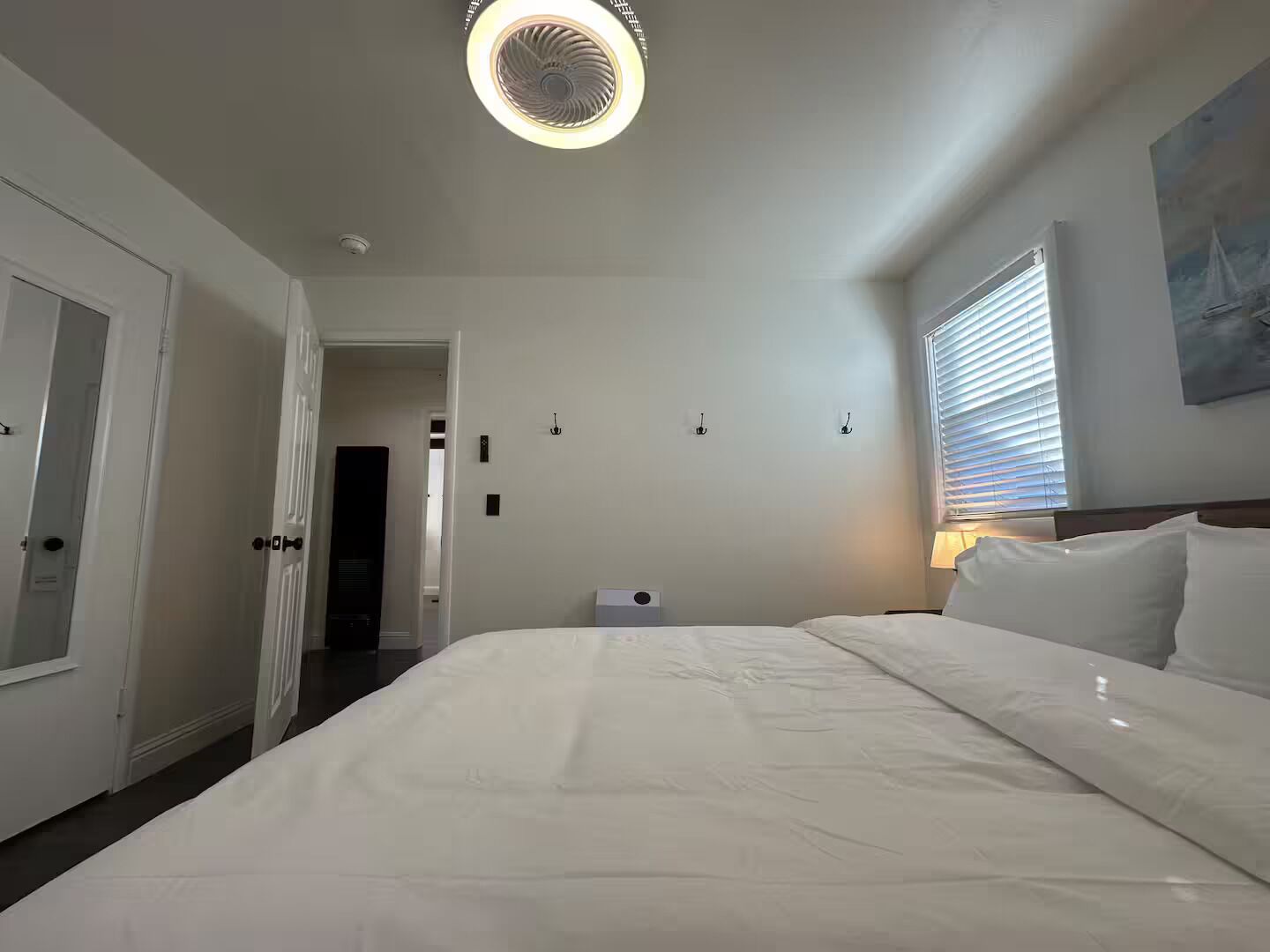 A hotel room with a large bed, a ceiling fan, a window with blinds, and a door leading to another room.