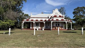 A white building with a red roof and a covered porch, surrounded by grass and trees. - Kalimna Homestead, Historic Stays, Hunter Valley (Paterson)