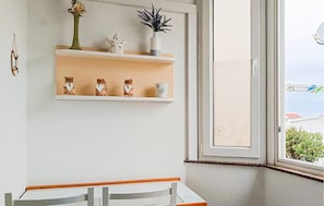 A room with a white wall, a window with a view of a house and greenery, a white shelf with decorative items, and a white table with a chair.