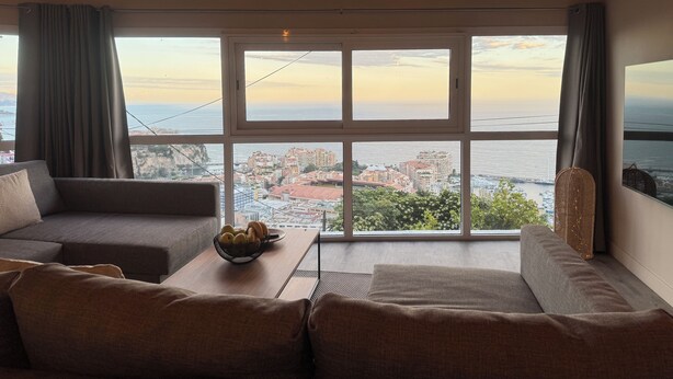 Sala de estar moderna com uma grande janela com vista para a cidade, um sofá, uma mesa de centro com uma tigela de frutas e um lustre.