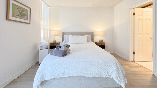 A neatly made bed with a white comforter and pillows, two bedside tables with lamps, a framed picture on the wall, and a door to the right. - #11 Modern 2-BR sanctuary walk to shops & beach (Santa Monica)