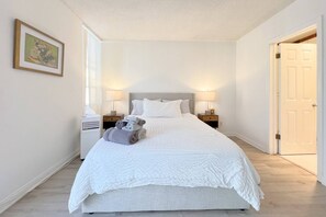 Ein ordentlich bezogenes Bett mit weißer Decke und Kissen, zwei Nachttische mit Lampen, ein gerahmtes Bild an der Wand und eine Tür rechts.