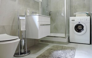 Ein modernes Badezimmer mit Toilette, einer in Glas abgeschlossenen Dusche, einer Waschmaschine und einem weißen Waschbecken mit Seifenspender.