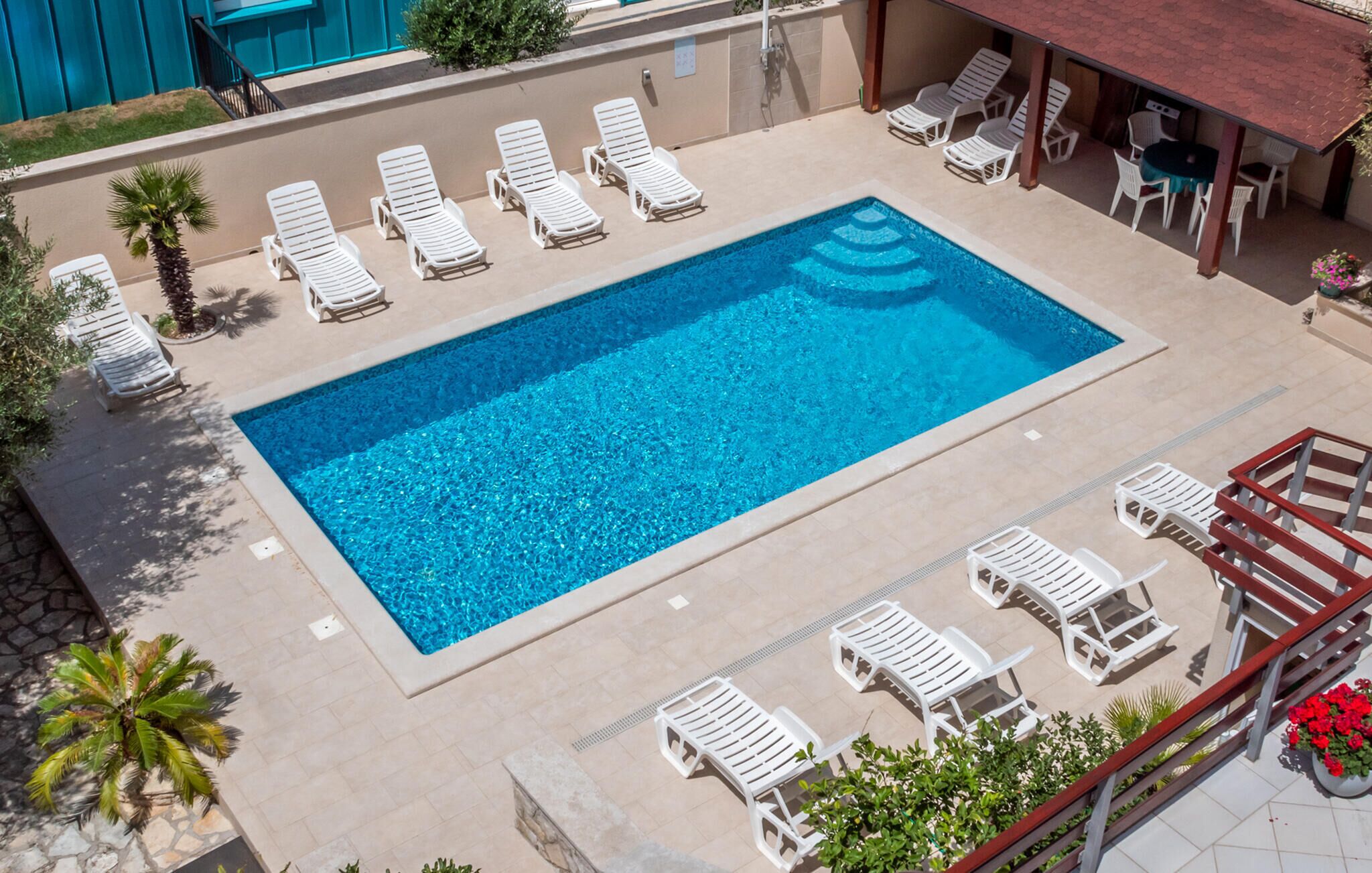 Ein rechteckiger Pool, umgeben von weißen Liegestühlen, mit einer überdachten Sitzecke und in der Nähe Palmen.