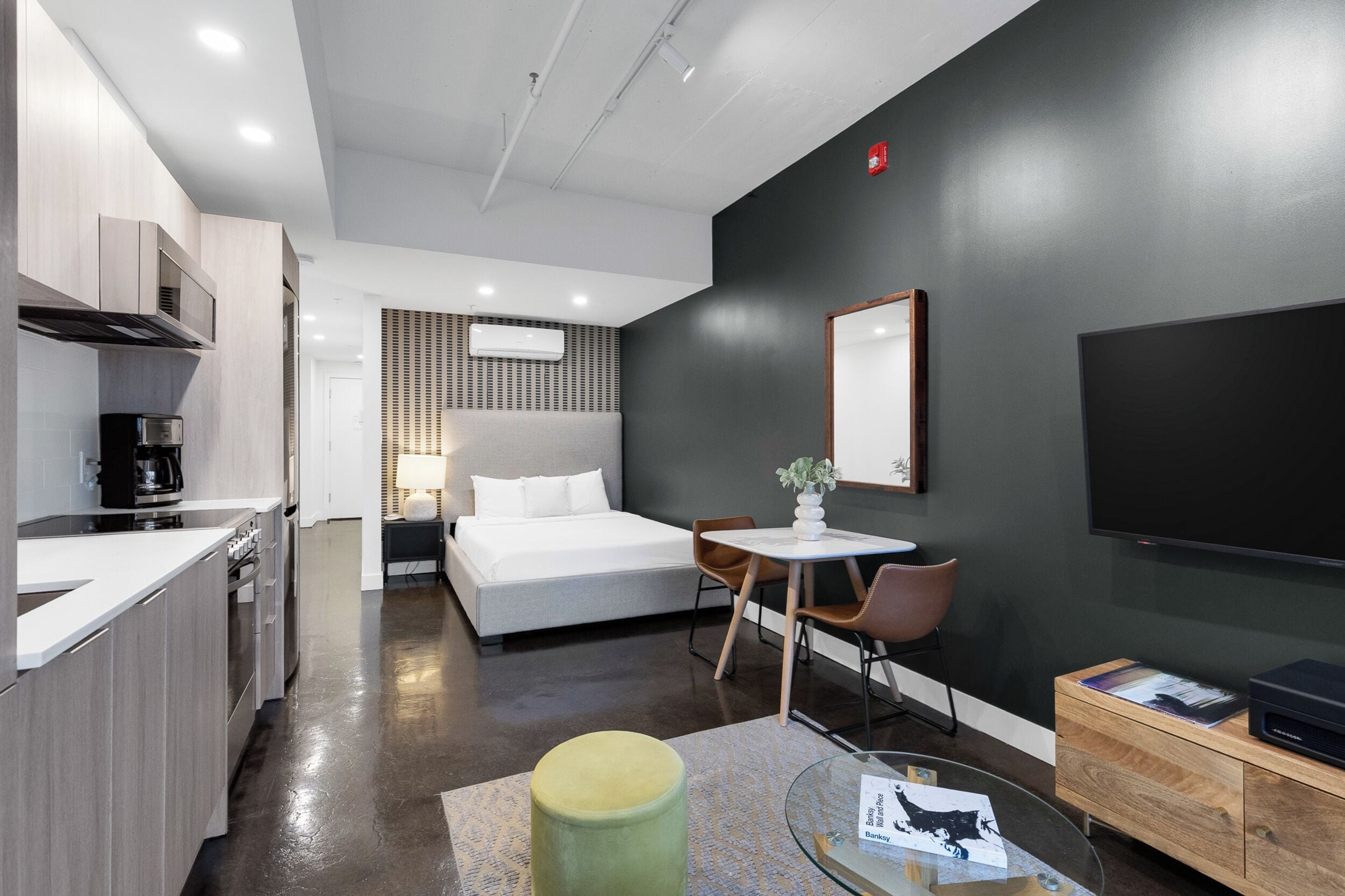 A modern hotel room with a kitchenette, a bed, a dining area, and a TV.