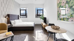 A modern hotel room with a bed, a desk, a chair, a TV, and a large window. - The Cove Hotel (Montréal)