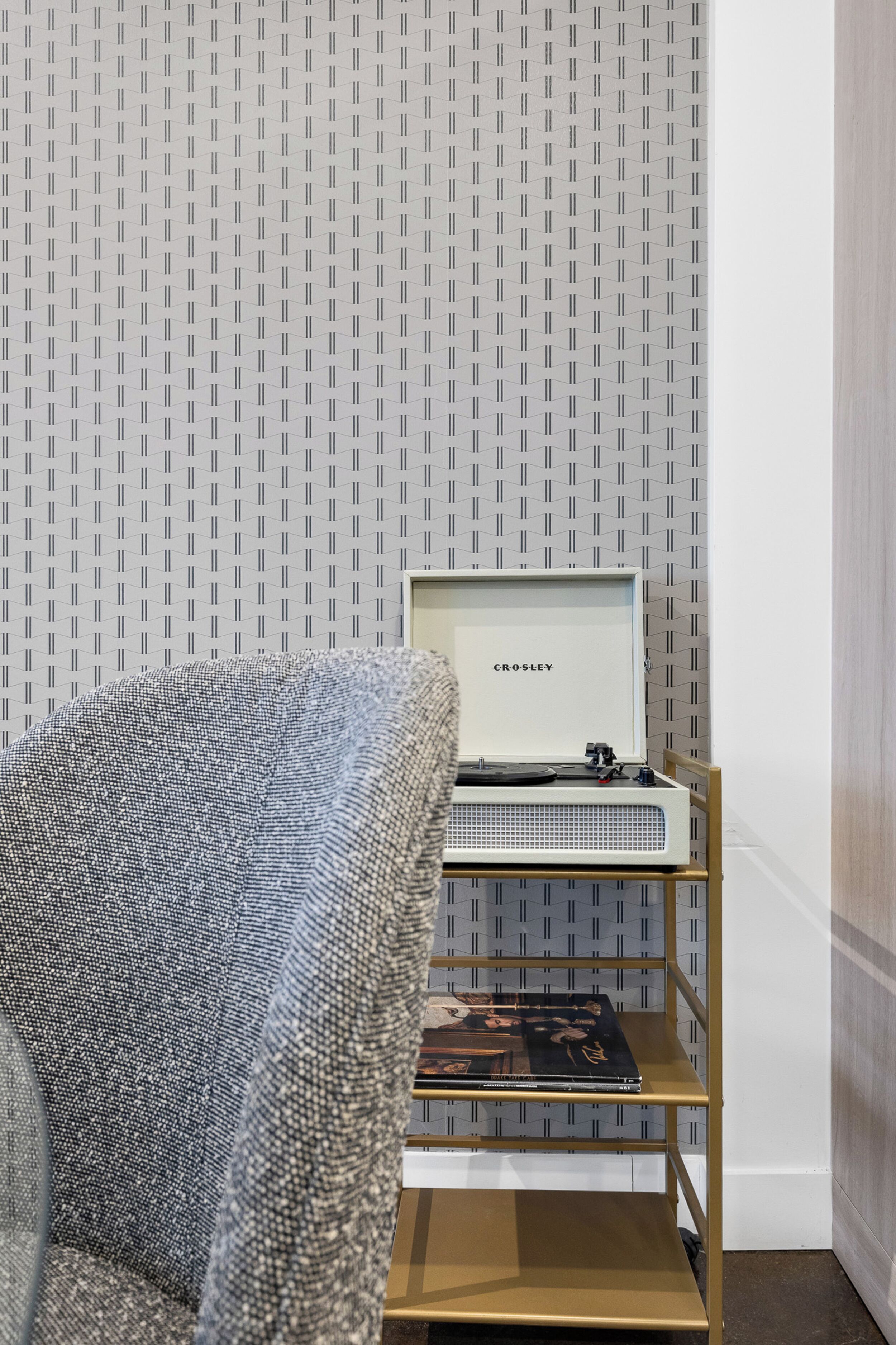 A room with a patterned wallpaper, a grey upholstered chair, a wooden shelf with a record player, and a framed item labeled "CROLEY" on the wall.