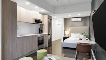 Cozinha moderna com eletrodomésticos de aço inoxidável, área de jantar com mesa de vidro e um quarto com cama e mesa de cabeceira.