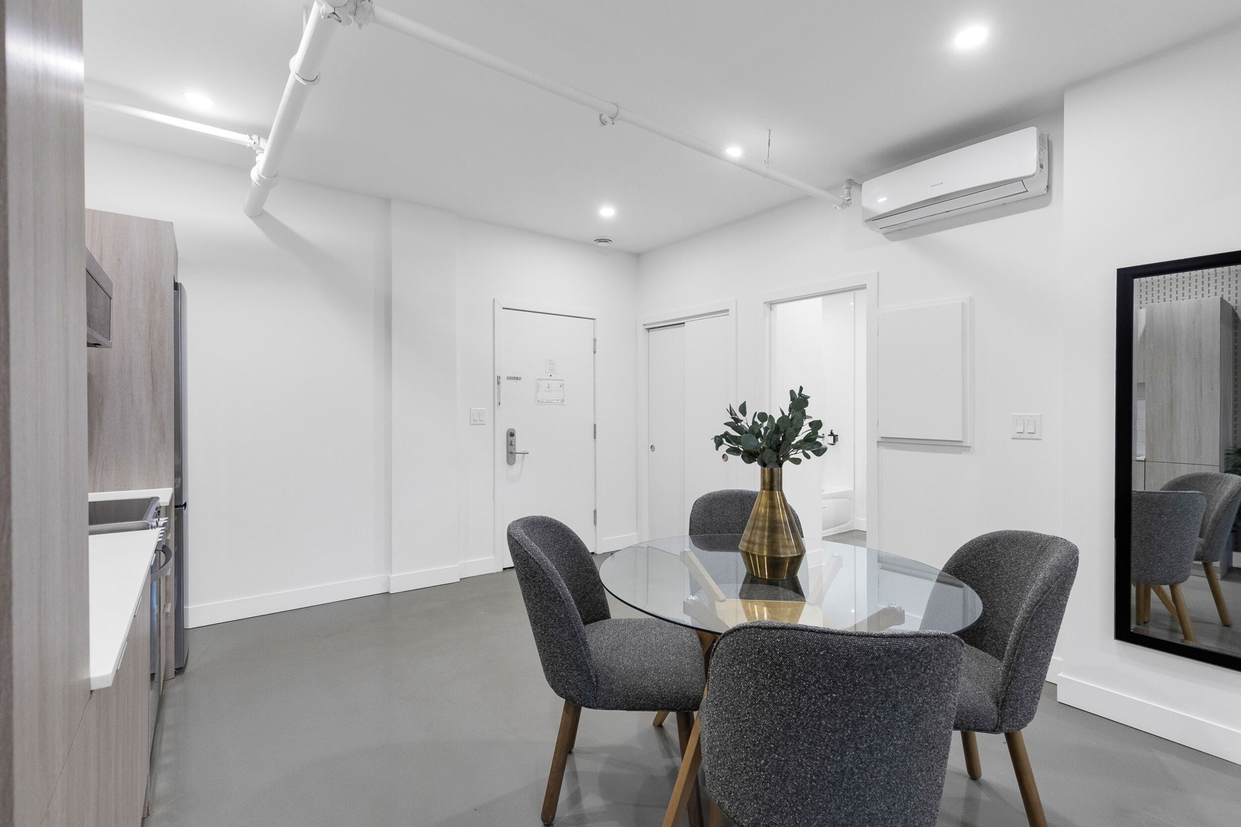 A modern conference room with a glass table, four grey chairs, a vase with greenery, and an air conditioning unit on the wall.