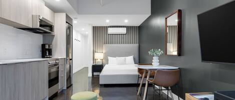 A modern hotel room with a kitchenette, a bed with white linens, a small round table with a chair, and a TV on the wall.