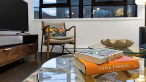 A modern living room with a glass coffee table, a television, a wicker chair, and two books titled 'The Catch' and 'The Hunter'. - The Cove Hotel (Montréal)