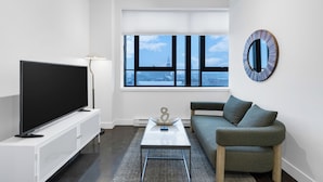 A modern living room with a large flat-screen TV, a coffee table, a sofa, and a window with a city view. - The Cove Hotel (Montréal)